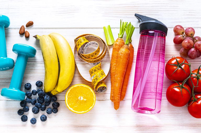 in order from left to right: two blue dumbbell weights, a bunch of blueberries, two bananas, a half of an orange facing up, yellow measuring tap, carrots, a pink water bottle, a bunch of purple grapes, and a vine of tomatoes 