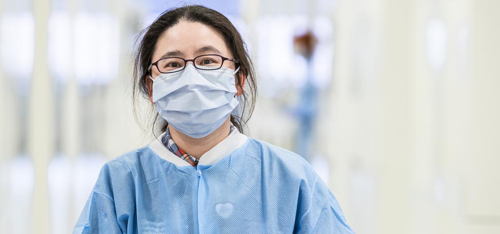 A woman with glasses, a blue medical mask, and blue scrubs faces the camera