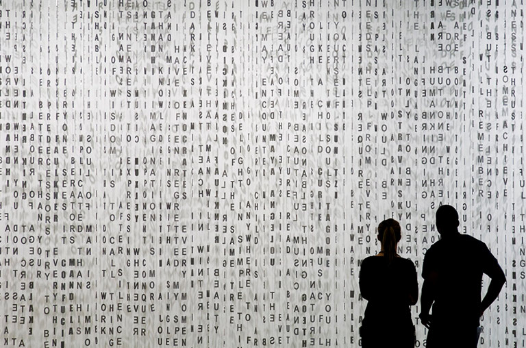 Image of two sillouthettes standing in front of an art piece of scrambled letters