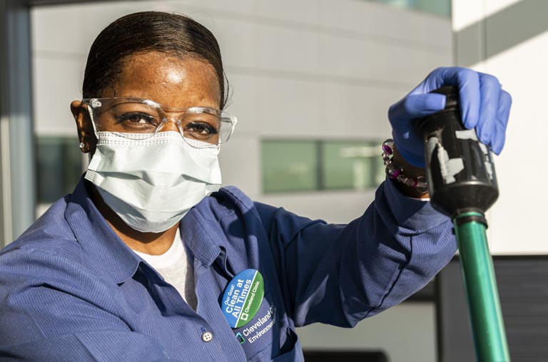Woman wearing glasses, a mask, blue Cleveland Clinic uniform, and blue gloves holding a medical instrument