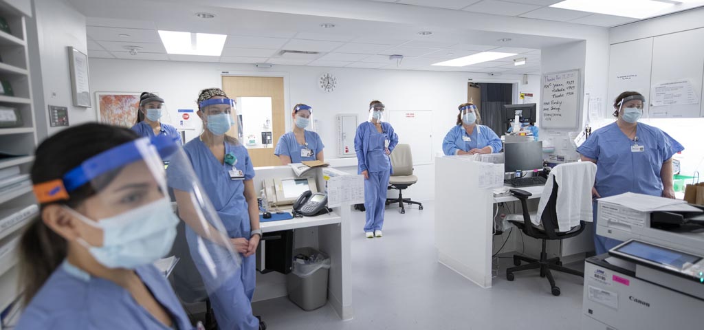 a group of eight people with a face shield, blue medical mask, and blue scrubs stand in a medical office room with desks