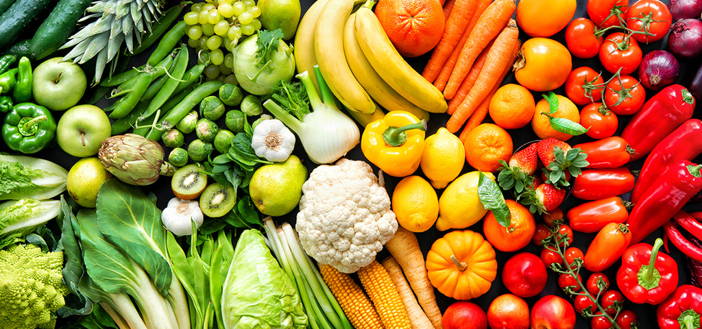 a grouping of fruits and vegetables organized by color. From left to right: a head of lettuce, cucumber, green bell peppers, granny smith apples, leeks, kiwis, brussels sprouts, garlic, cauliflower, bananas, yellow bell peppers, carrots, oranges, red bell peppers, tomatoes, strawberries, eggplant, grapes