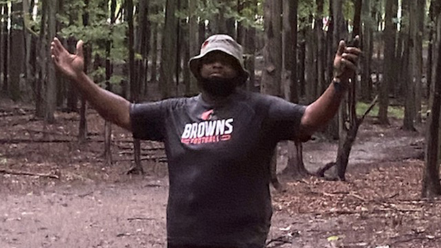 Man wearing a Brown's shirt and a bucket hat, with his arms up, in the woods.